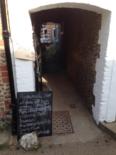 View through a narrow stone archway, down a lane. Book sale at the bottom of the lane. Signs in the foreground advertise used book sale and homemade jam for sale.